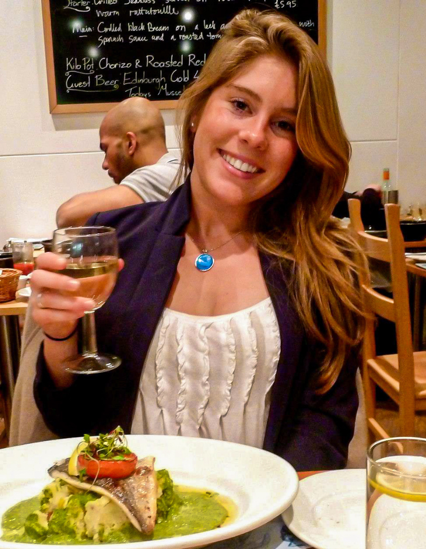 Headshot of Victoria Pardo, Full color, posh dining room in background, elegant meal in foreground and white wine glass in hand. 