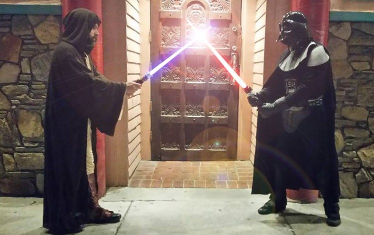 Mental Suppository hosts Bret Herholz and Andrew SHanley dressed in Star Wars garb in front of a dark stained ornate and large paneled door, light sabres touching and emitting light.