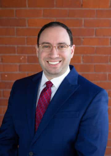 Gardner (MA) assistant city solicitor  Ethan Kolodny, Blue suit with red tie, white shirt with a brick wall behind him. 