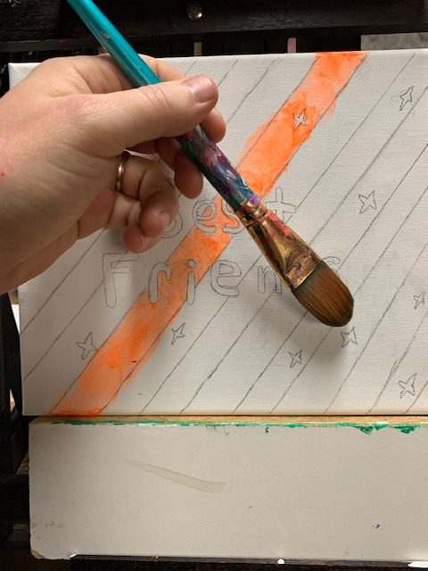 Paint Canvas Descriptive image white canvas with orange stripes, in process of being painted with word best friends in the center. hand coming into the image from the left holding a blue artist's paint Bruch in position to paint the canvas.