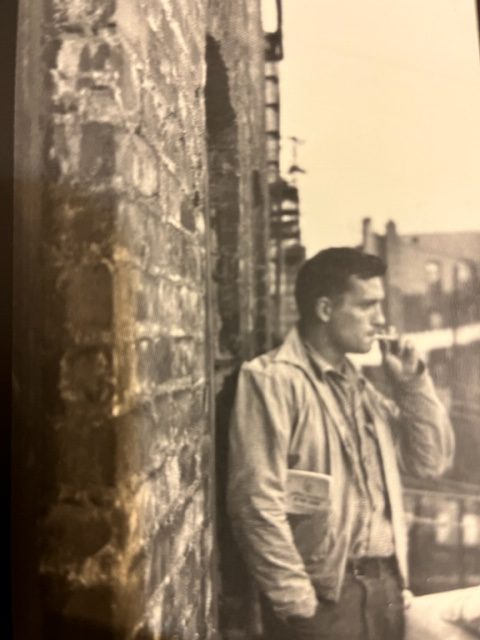 Historic photo of Jack Kerouac, the Beat Generation author, black and white, mid-20th century, leaning on a brick wall and smoking a cigarette