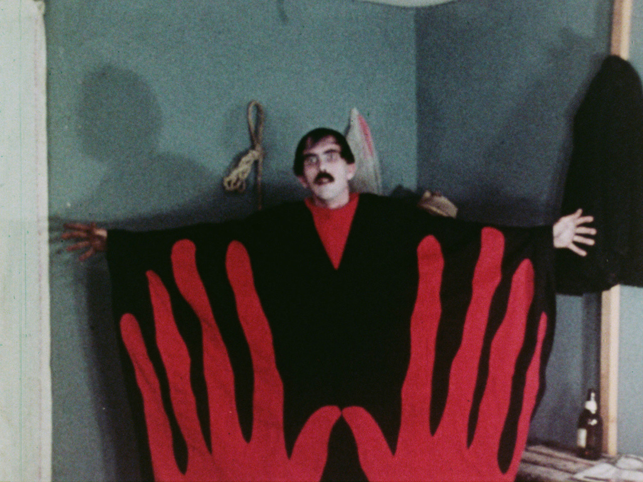 Full Color Image from the Z movie Manos Hands of Fate.  Black-haired man in white face paint wearing a black on red aboriginal-like robe, hands outspread across from the shoulder