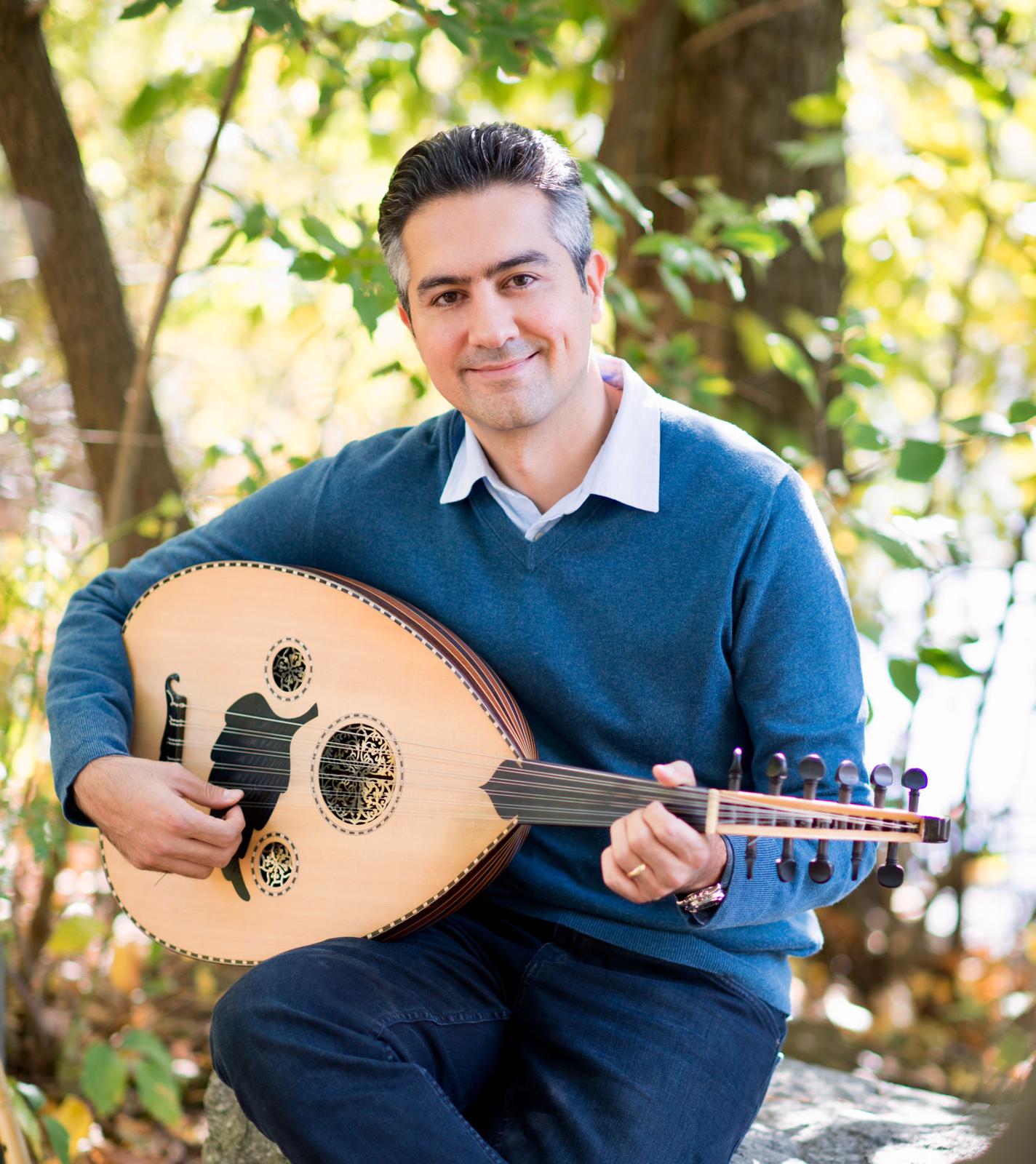 Outdoor full body image of Musician Volkan Efe.  Sitting down in wooded backdrop with a Turkish stringed instrument known as an Ud.  Blue sweater, white shirt.  Blue slacks and smiling. 
