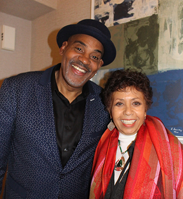 Full color photo of Jazz Guitarist Allan Harris standing stage left to show host Joan Watson-Jones.  He's is in a blue suit coat, black fedora and black Oxford shirt.  Joan is wearing a red silk scarf over a dark suit. 