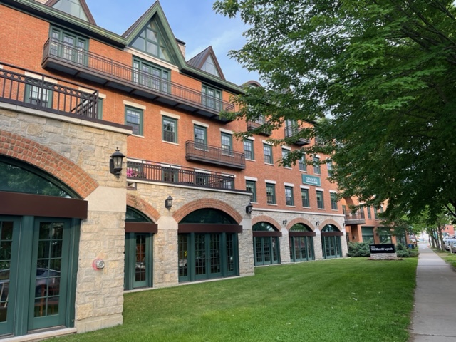 Full color image taken in the evening of the development known as Main St. Landing in Burlington, VT. Brick and stone facade on a tree lined street. Green Lawn in a strip between sidewalk and building front.