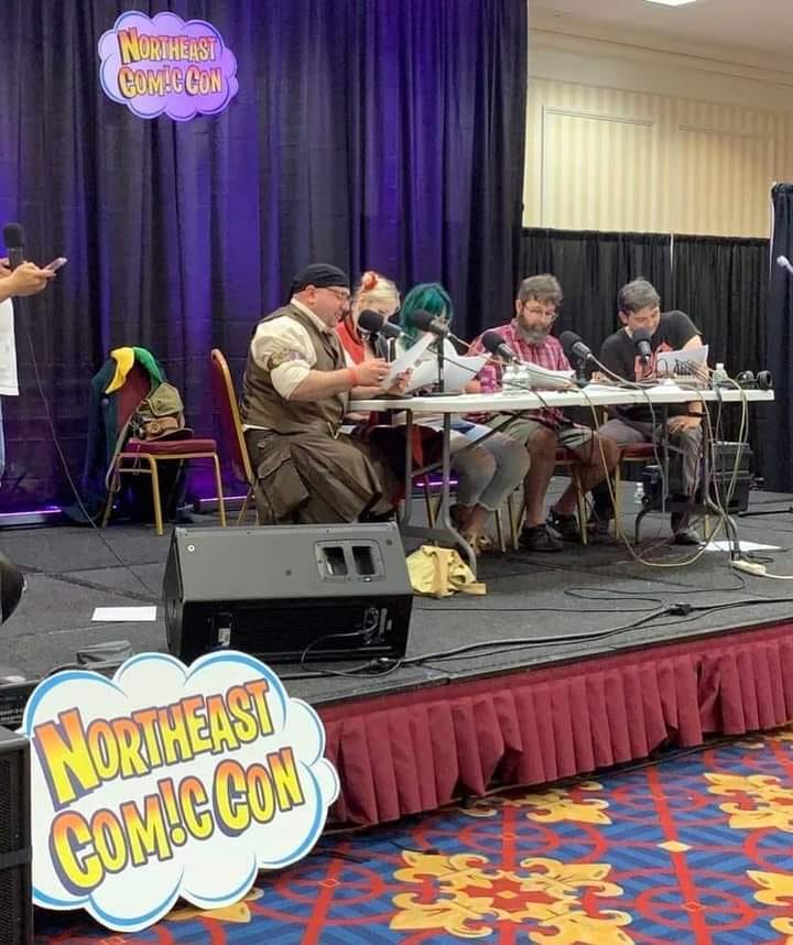 An image of mental suppository cast members sitting at a table in front of a live audience to read a script during Northeast Comic Con. Large sign in hot colors to the left of stage and purple curtained background in a hotel convention room