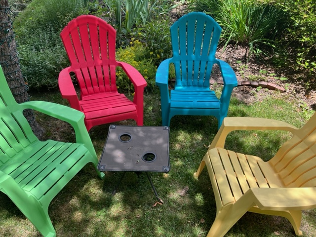 Color image of four plastic Adirondack chairs, from left to right green, red, blue, yellow and surrounding a black table.  dappled sunlight covers the area in a scenic backyard.  Symbolic of DEI.