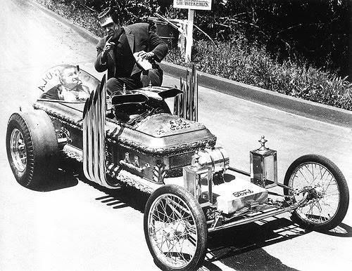 Black and White mid-1960's image of 'Dragula', a hot rod modified once used on the hit Television series, 'The Munsters'. Several characters from the show stand aside or sit in the vehicle.