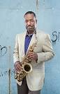 Donald Harrison Alto Saxop[hone in hand, DOnald Harrison is wearing a white sport coat and purple shirt, headshot against the backdrop of an urban wall background.