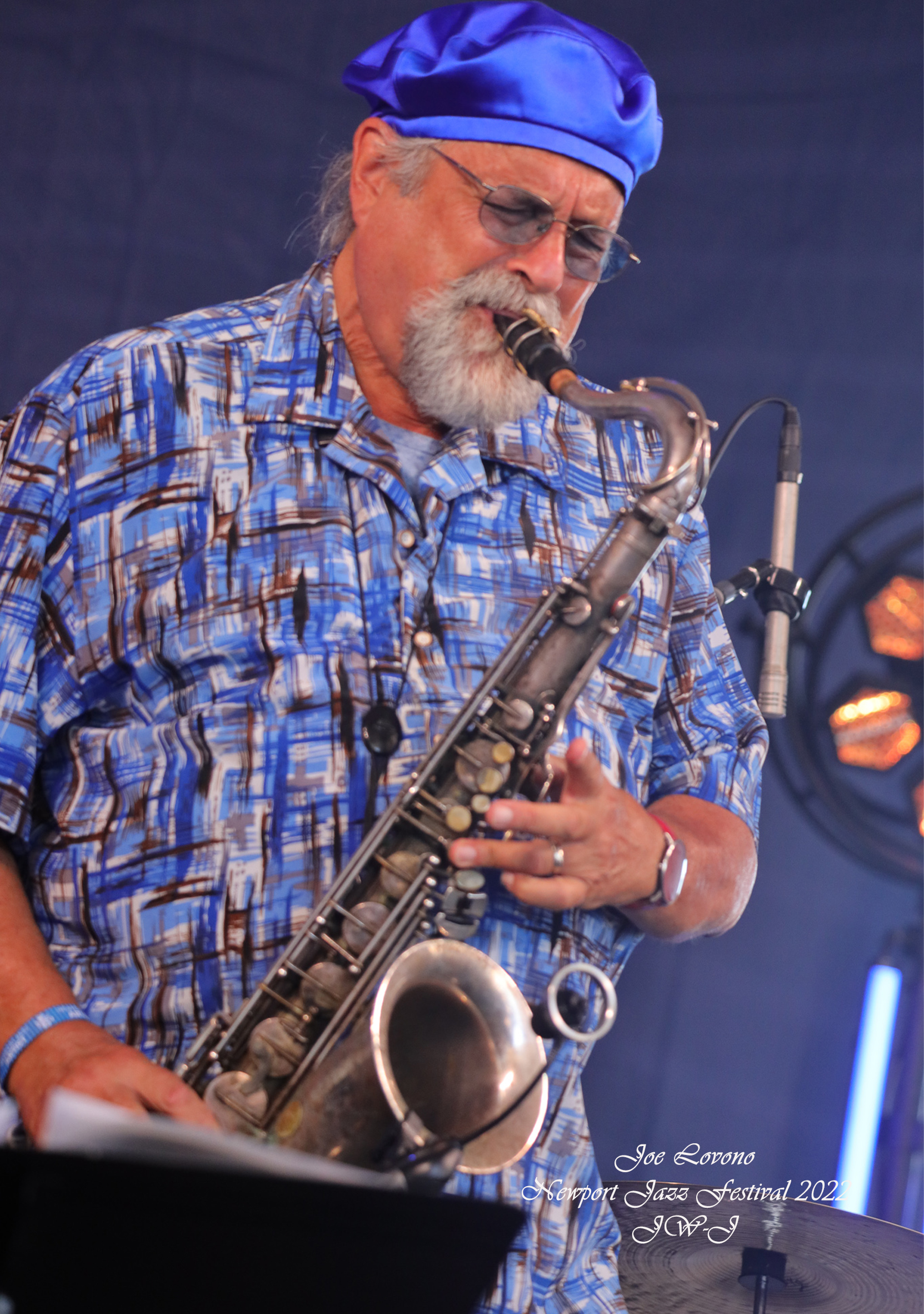 Full color image from Newport Jazz Festival 2022 of Tenor Saxophonist Joe LOvano on-stage with his horn in play, blue satin beret and colorful Oxford shirt. 