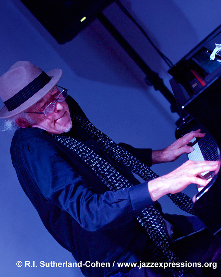 PIanist and Magazine Publisher David Haney, standing while playing a piano, dark blue suit, grey fadora hat on his head, dark blue background