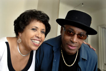 Full color image of R&B-infused Jazz Vocalist Lee Genesis (right) with The Jazz Room host Joan Watson-Jones.  Lee wears a black porkpie hat, blue shirt over black tee and gold chain.  Joan is smiling and wearing white with solver earrings.