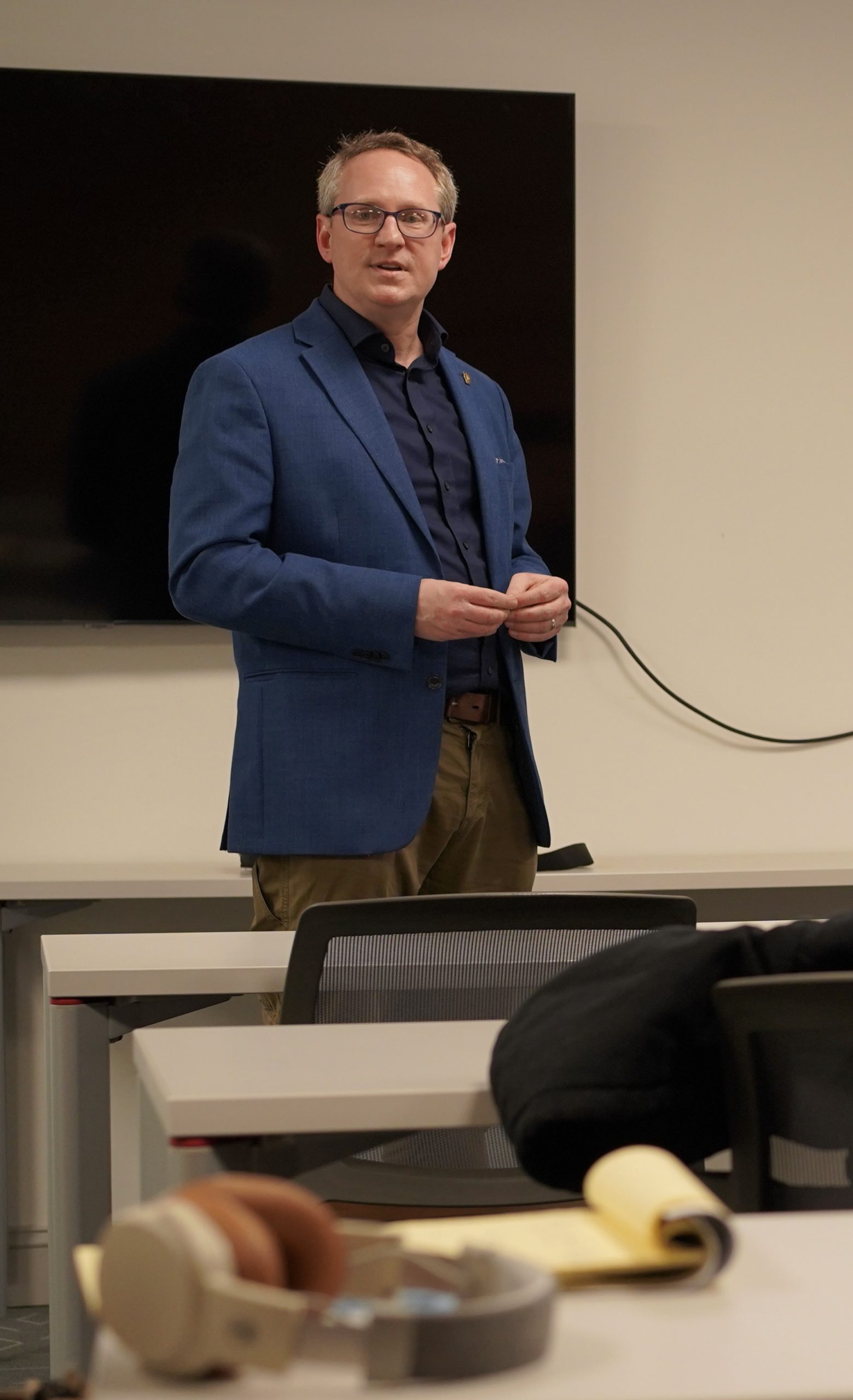 Full color image of Director Scott M. Graves, Startup Rutland presenting during a recent class on business development topics. Blue soprtcoat and shirt, a man wearing glasses, hands come together and speaking to a crowd of adult students.