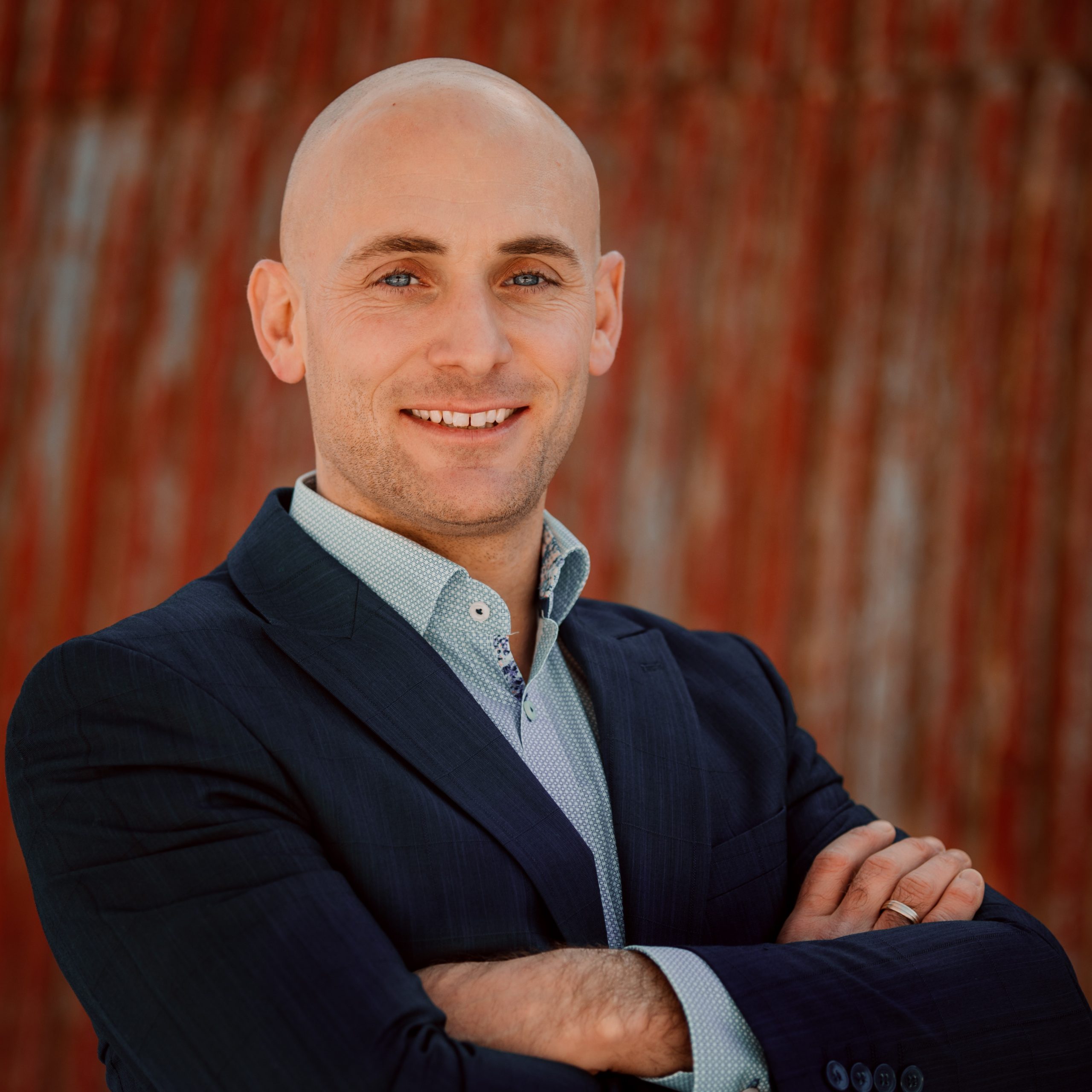 Full color headshot of Nattie Hussey, Economic Development Professional.  Blue sport coat, arms folded with an industrial sheetmetal background