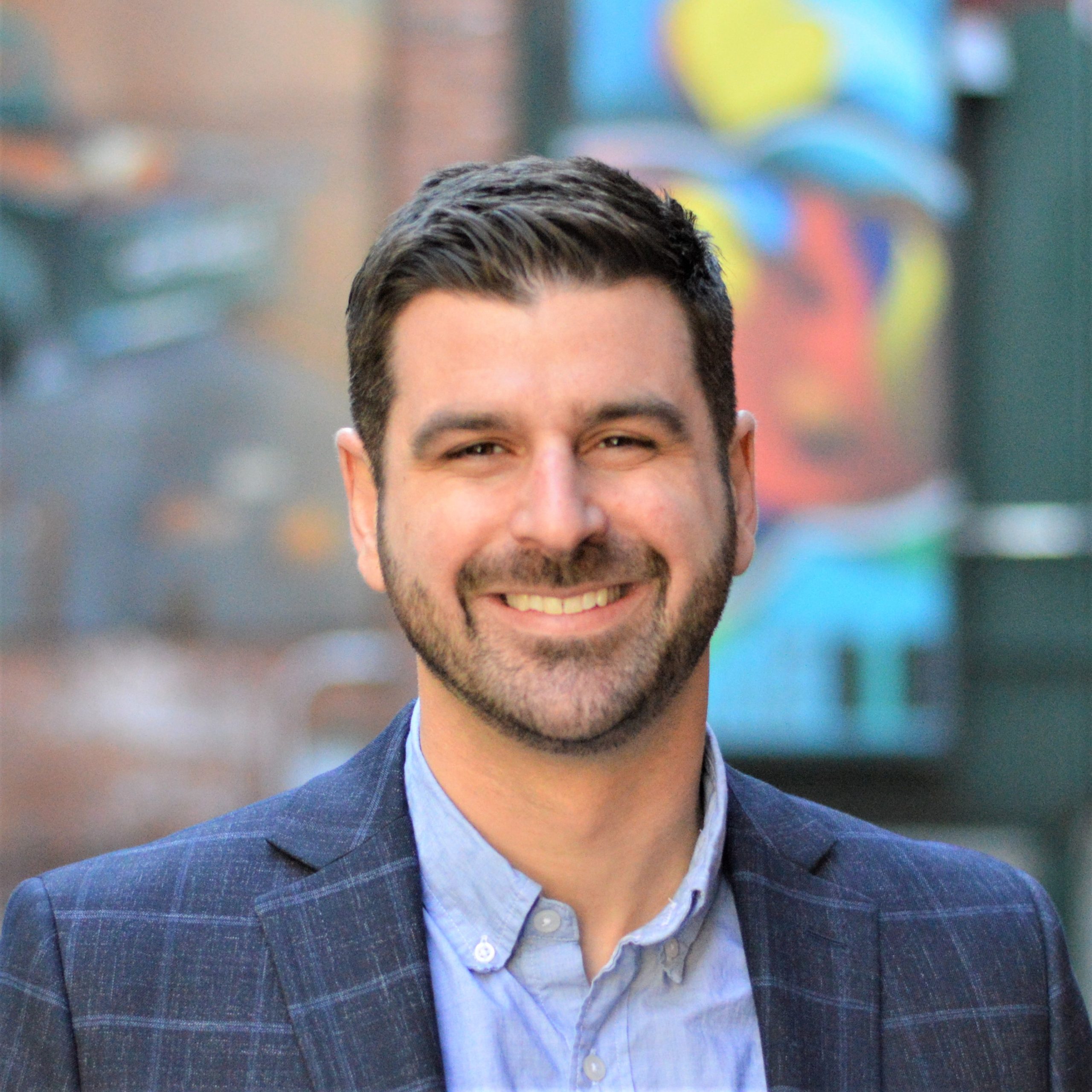 Full color headshot, blue sport coat and shirt, urban brightly colored exterior background of our guest Devon Neary